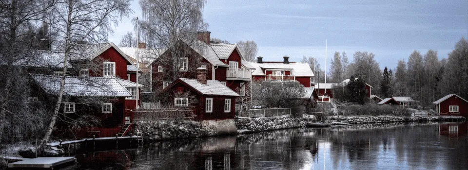 Traditional red wooden houses by a calm river in winter landscape, reflecting Missing Sweden While Being Abroad and longing for familiar Nordic scenery.